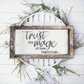 Wood-framed Trust Magic sign styled with greenery on a white wall.