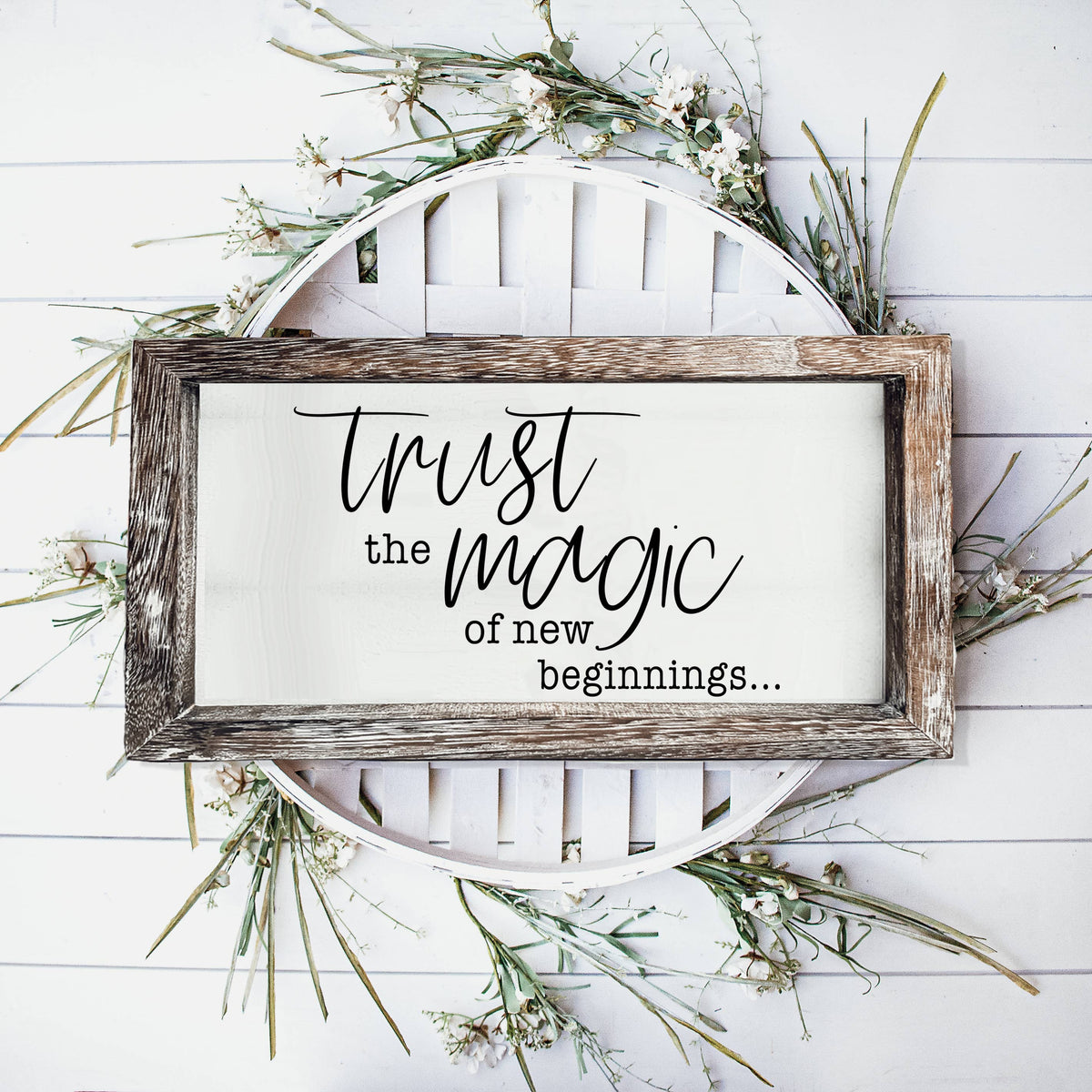 Wood-framed Trust Magic sign styled with greenery on a white wall.