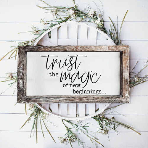Wood-framed Trust Magic sign styled with greenery on a white wall.